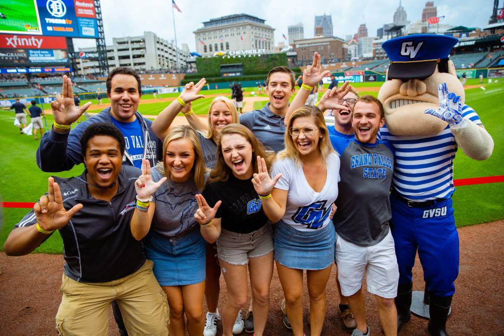 A group of gsvu choir students are standing on the infield of comerica park with louie the laker and everyone is holding up the gvsu 'anchor up'